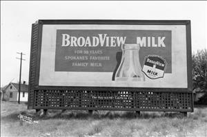 A billboard in a grassy field shows a picture of a glass milk bottle and the words Broadview Milk. There is a white house in the background
